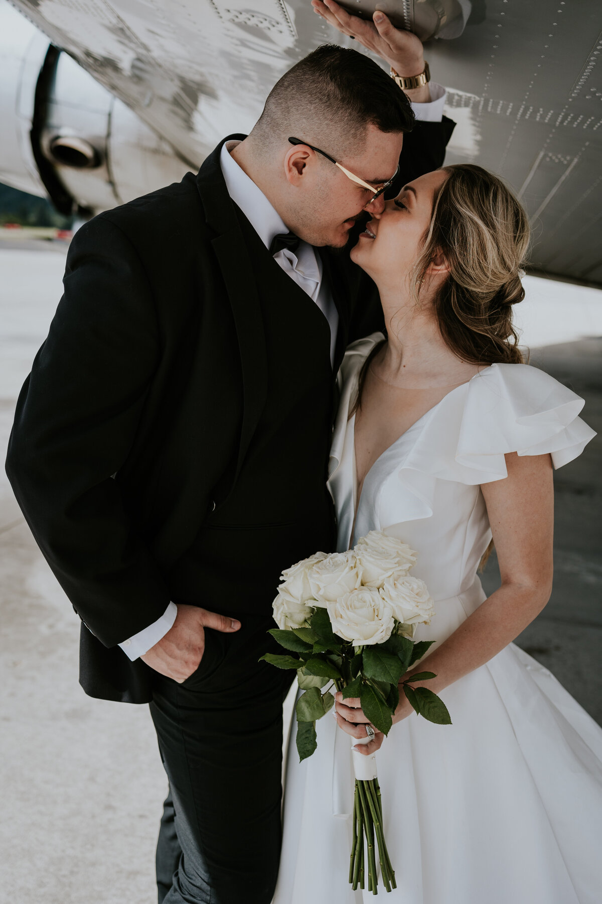 Groom leans in to kiss bride while standing underneath WWII plane wing.