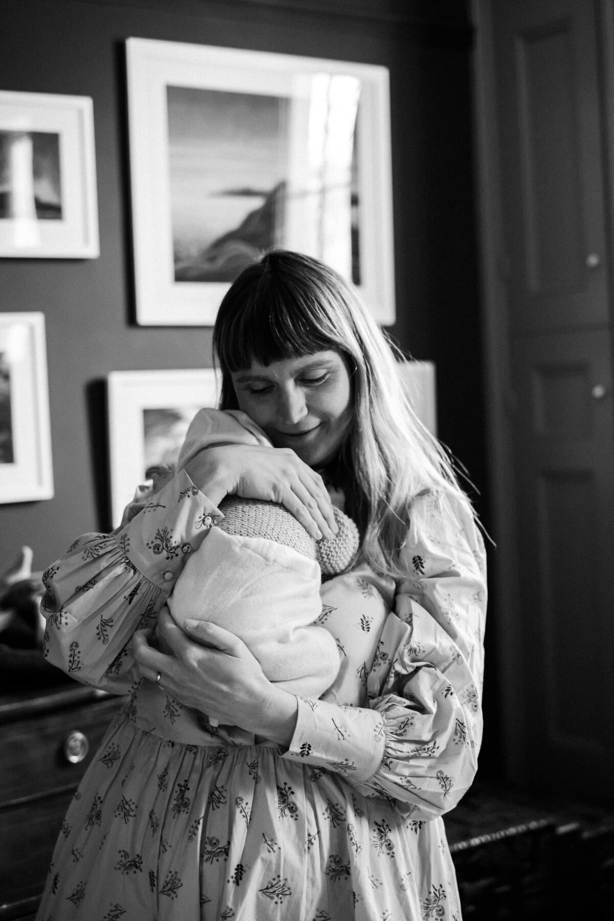 Mum cuddles her newborn baby standing in front of wall full of framed artwork