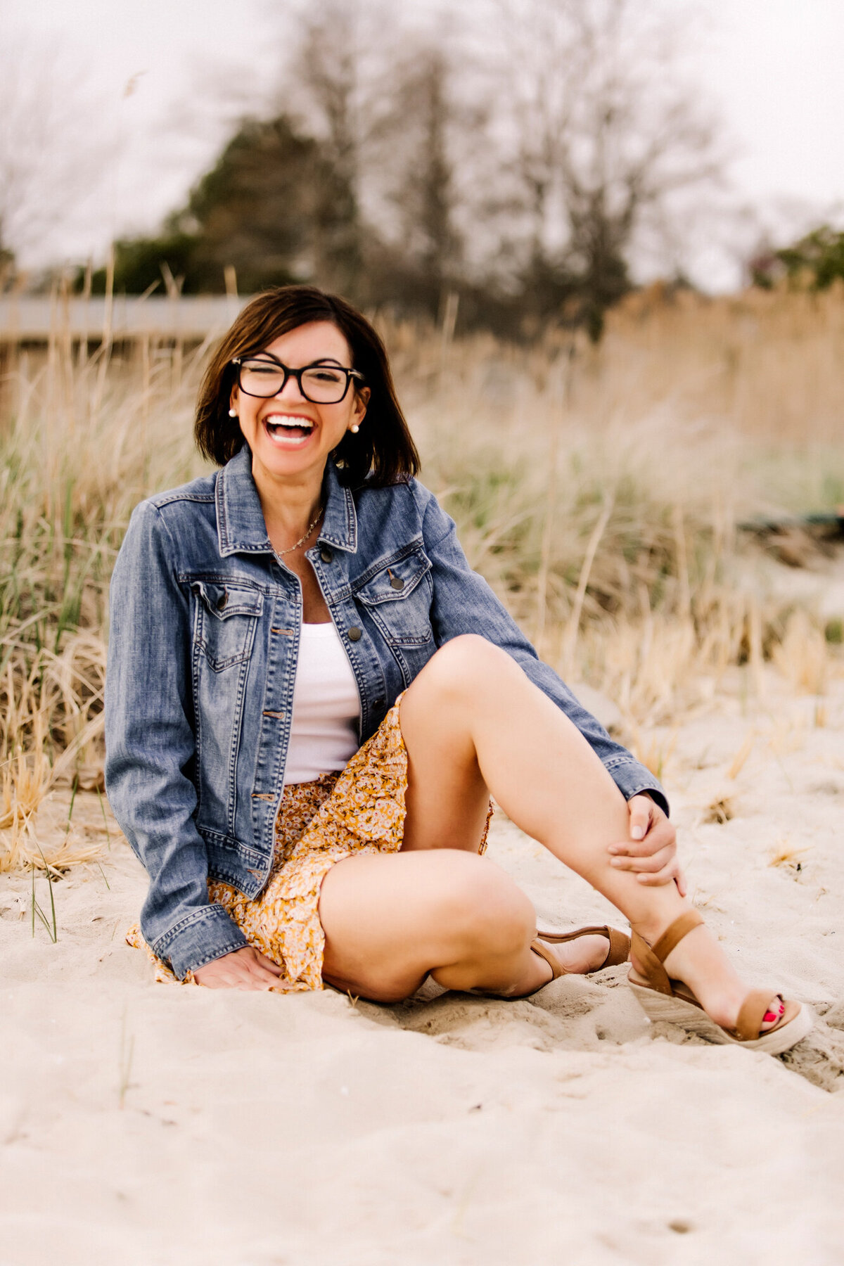 A female dating portrait client wearing a jean jacket over a white shirt & flowy yellow skirt, sitting on the sand while laughing
