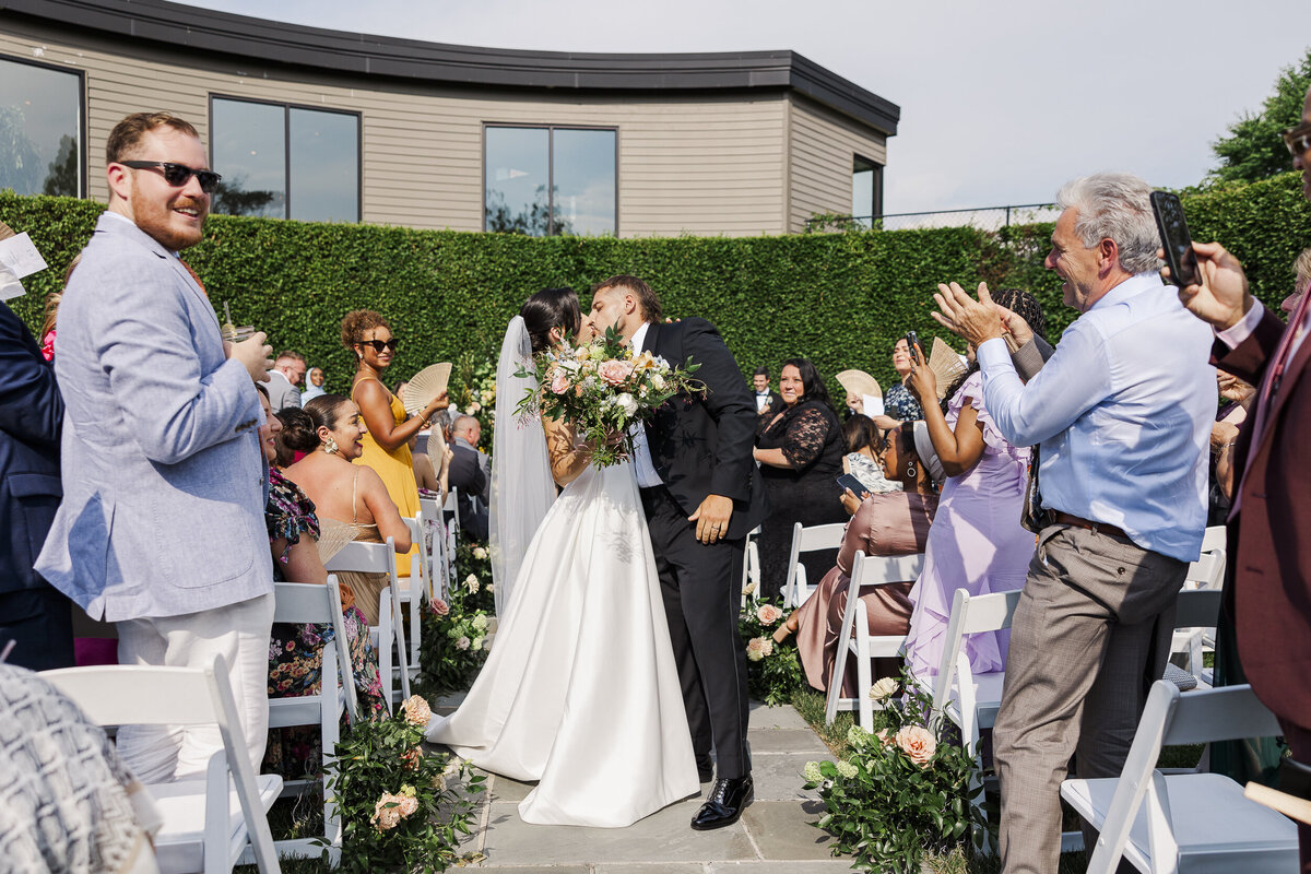 Bride and groom go for a kiss in the middle of the aisle during their outdoor ceremony at The Roundhouse
