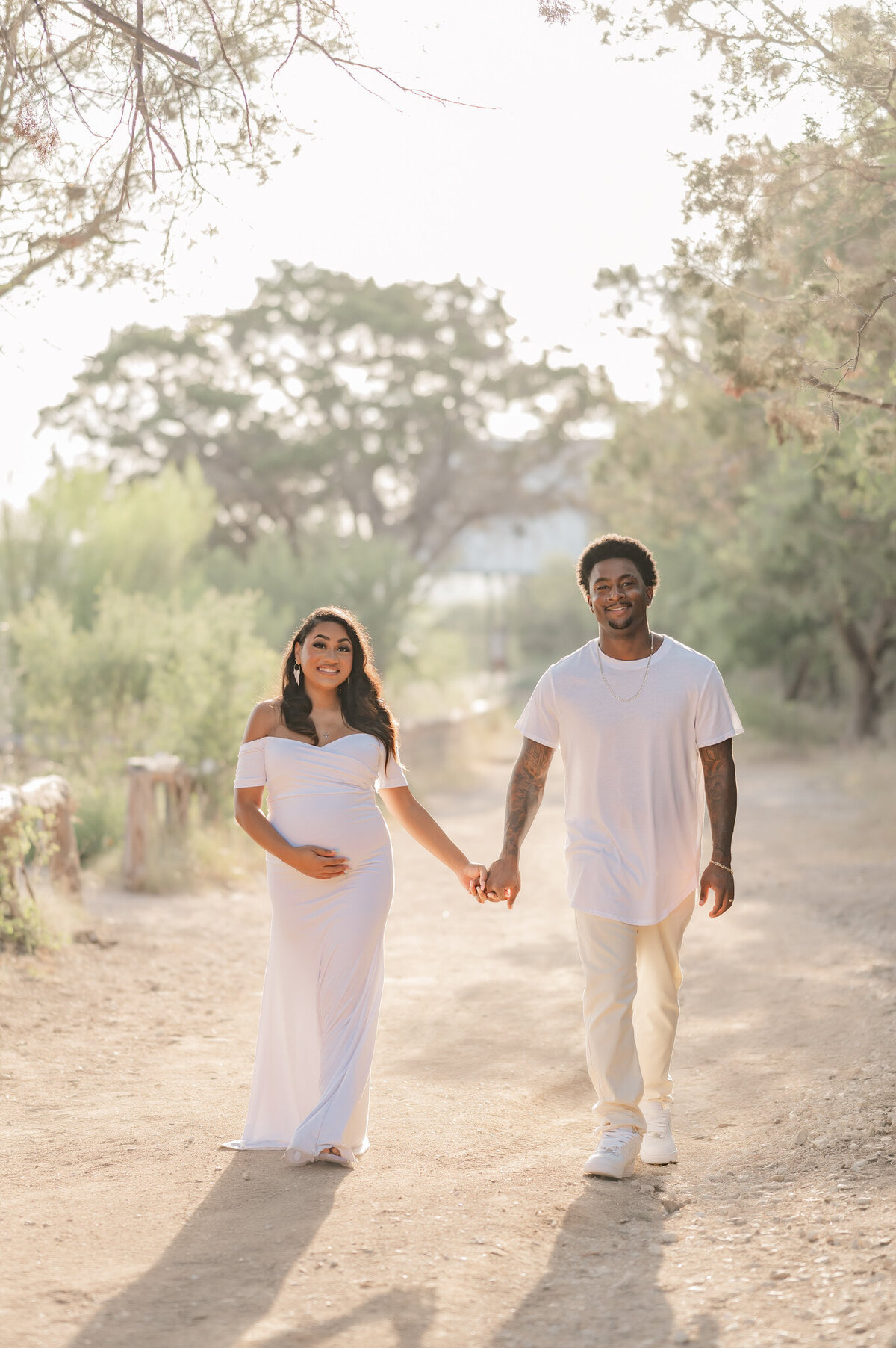 Young expecting couple in all white walks towards the camera holding hands.