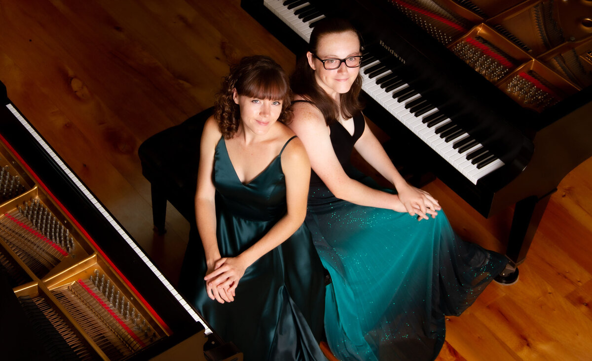 Classical music duo portrait Kaitlin and Emily Webster Zuber sitting backs against each other wearing green dresses amidst pianos