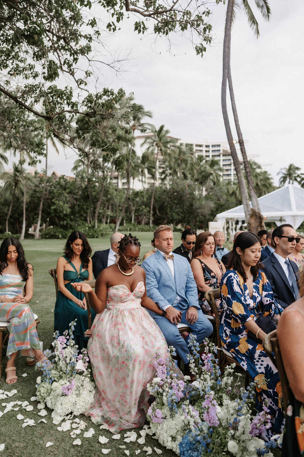 wedding guests closing their eyes for prayer at lanikuhonua ceremony surrounded by light lavendar, blue ane white florals
