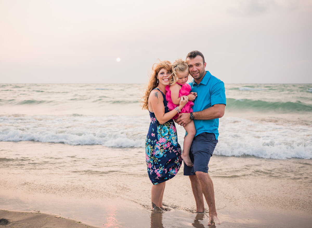 A family of three posing together on the beach at sunset, with the waves gently touching their feet