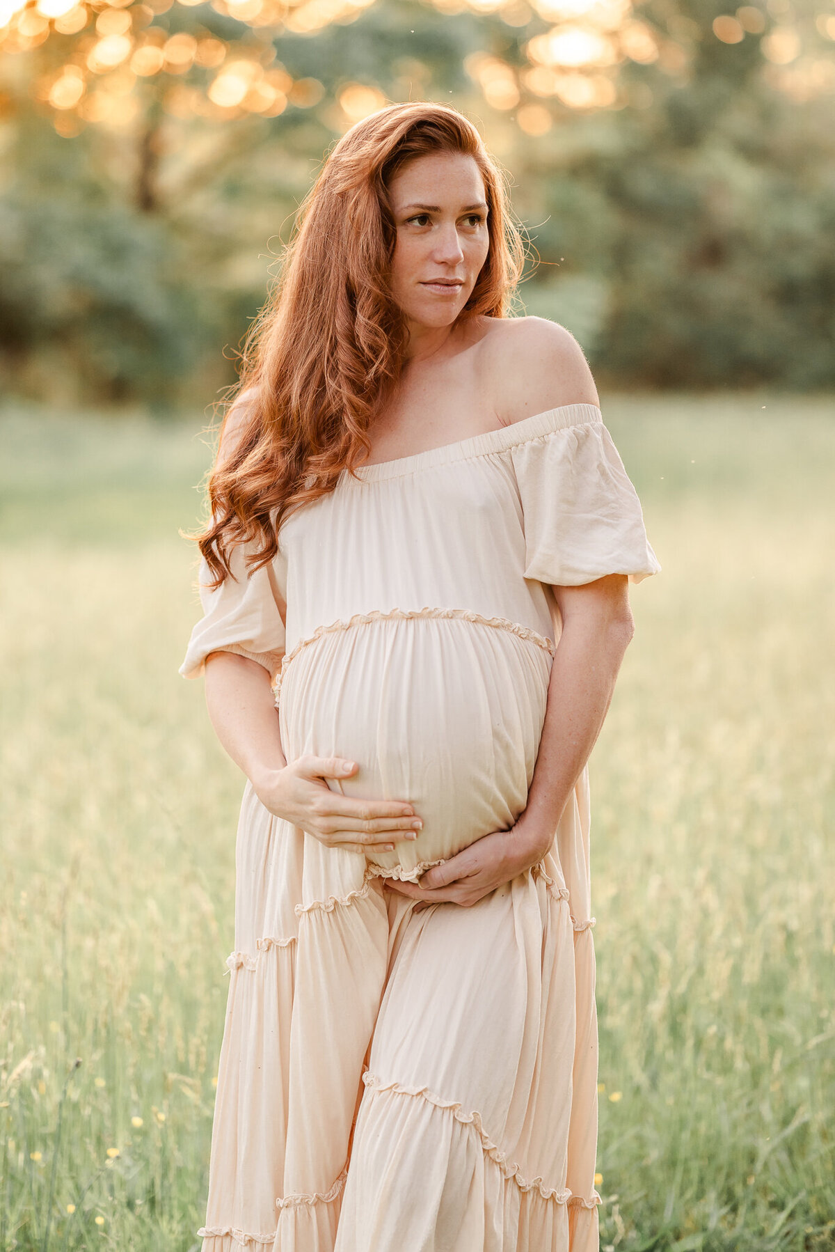 A mama-to-be, wearing an off-white simple gown, stands in a grassy field. She is holding onto her baby bump.