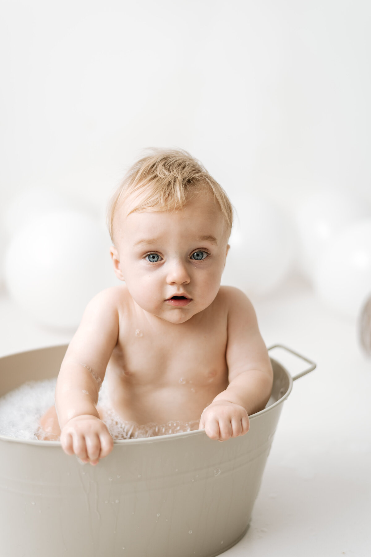 Baby boy sitting in a bath tub holding onto the side at cake smash photoshoot in Billingshurst