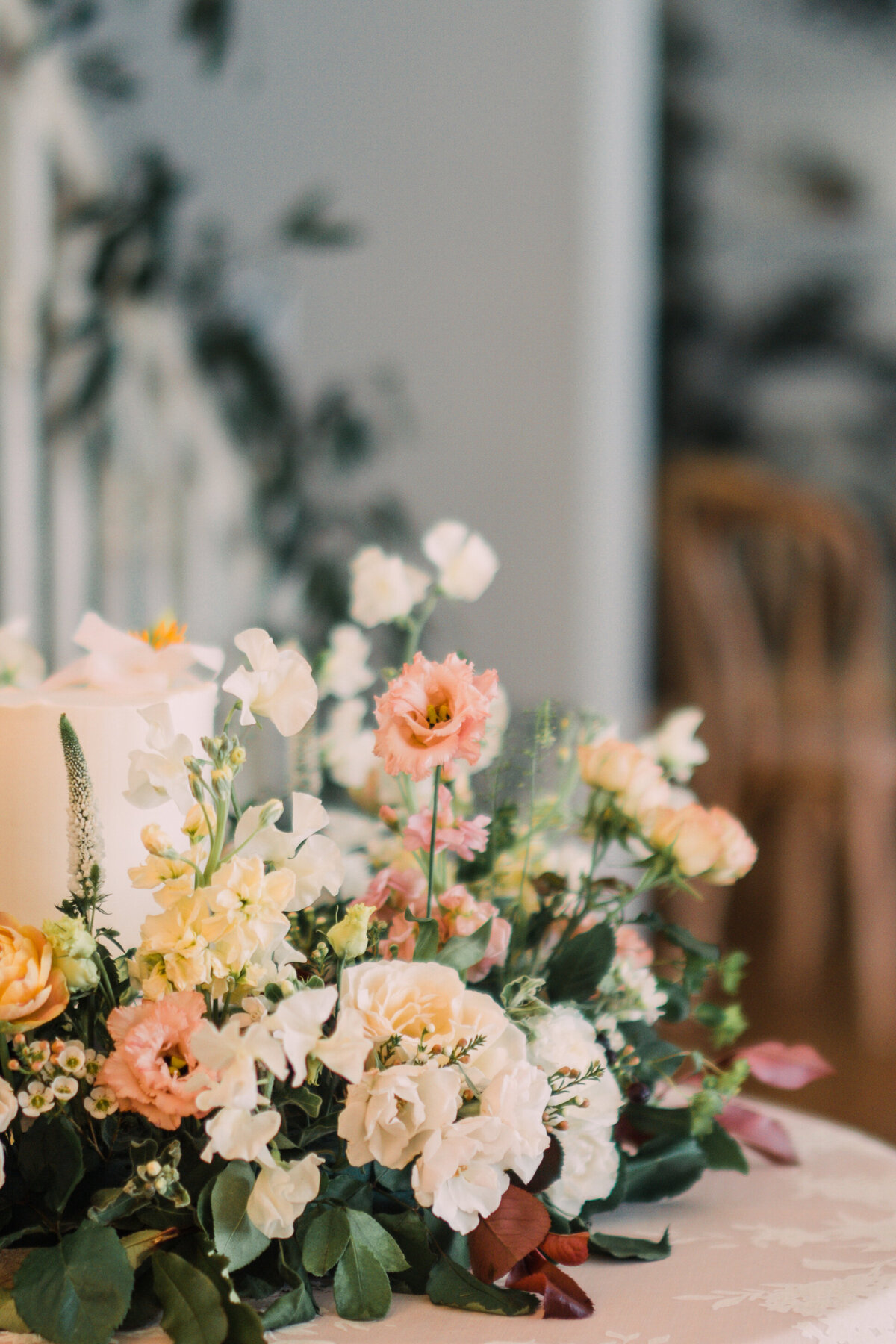 Closeup of wreath of roses, lisianthus, greenery, and red foliage around a cake with vines and chairs in the background