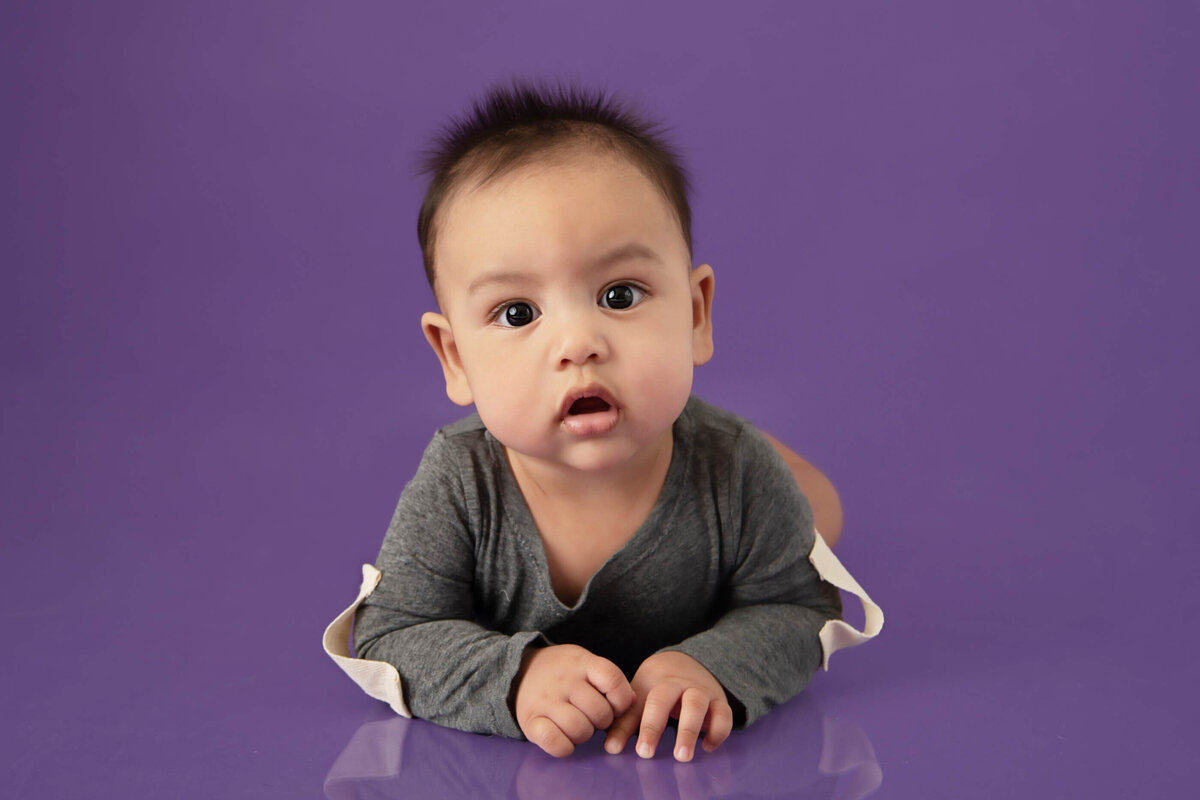 Baby boy wearing a grey onesie laying on his stomach on a purple background for his kids photography session in Burlington, On