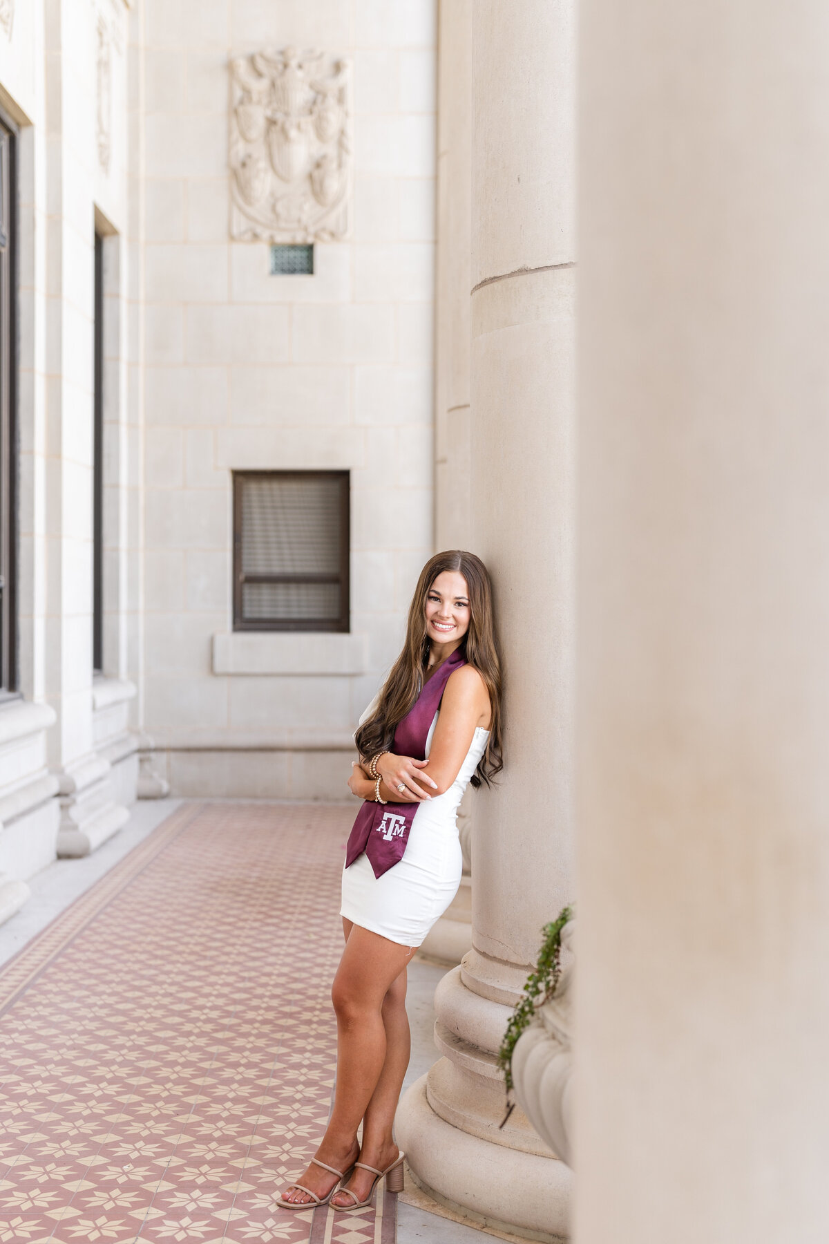 Texas A&M senior girl leaning against column and crossing arms while smiling and wearing white dress and maroon stole at Administration Building