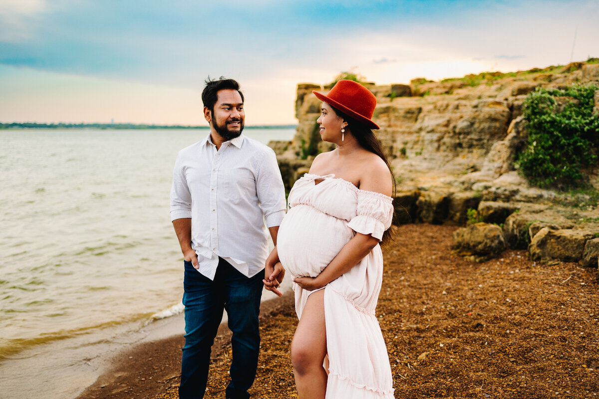 Photo of a pregnant couple in front of the sea, the woman has a red hat and a long white dress and is looking at the face of the man who is smiling in a white shirt and jeans