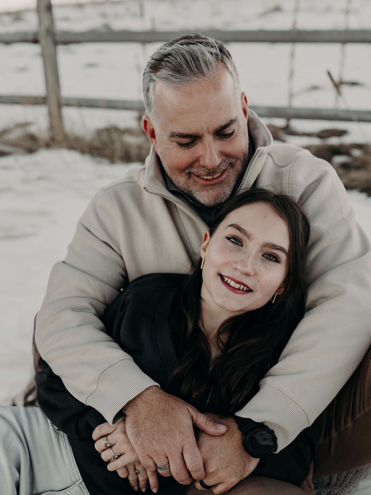 A teenage daughter, lovingly lays cuddling with her dad during an outdoor snowy photography session. She almost looks like snow, white with her beautiful, fair skin and red lips, while the father has salt and Pepper colored hair and is wearing a cream colored sweater.