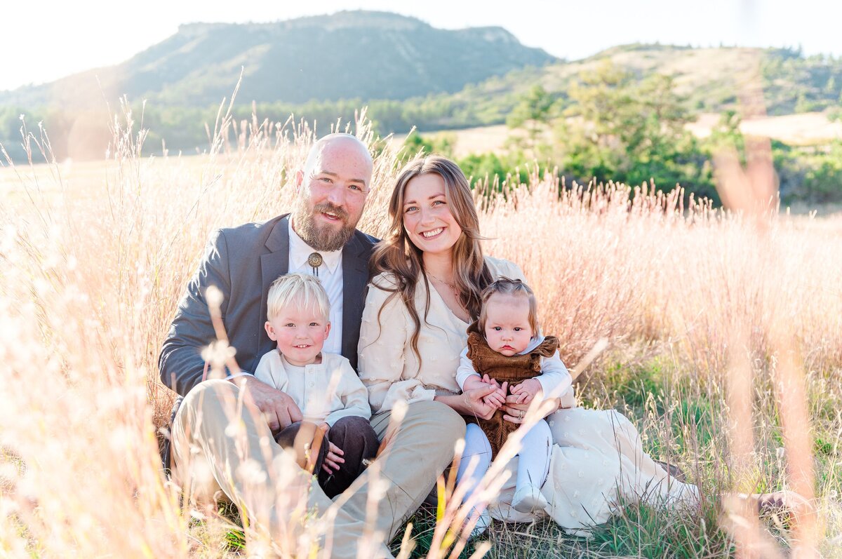 A couple in neutral tan and gray outfits sit on the grass field in front of a range of mountains captured by Denver family photographer