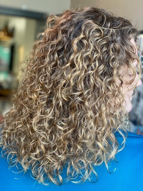 A woman with shoulder-length, naturally curly hair, showcasing the beautiful and defined curls styled by 212 Salon, Spa, & Barbershop.