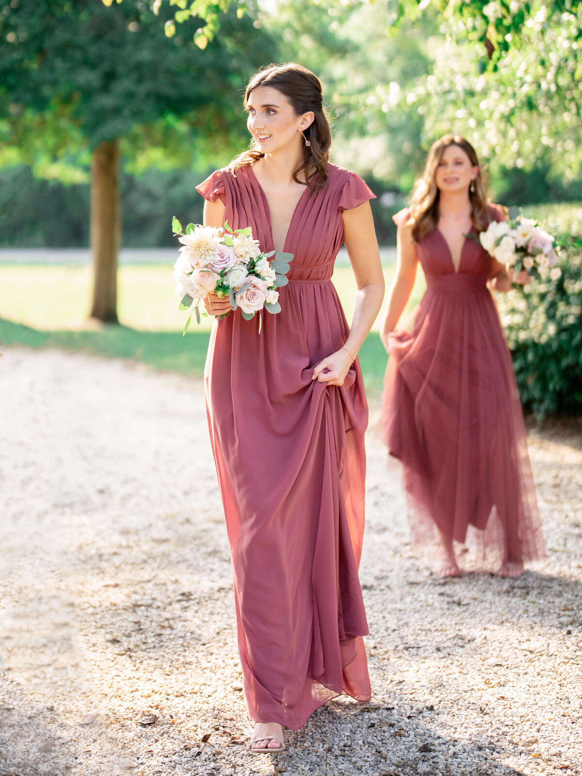 Two women in flowing mauve dresses walk outdoors on a gravel path. Each holds a bouquet of white and pink flowers. Trees and greenery create a lush background under a sunny sky.