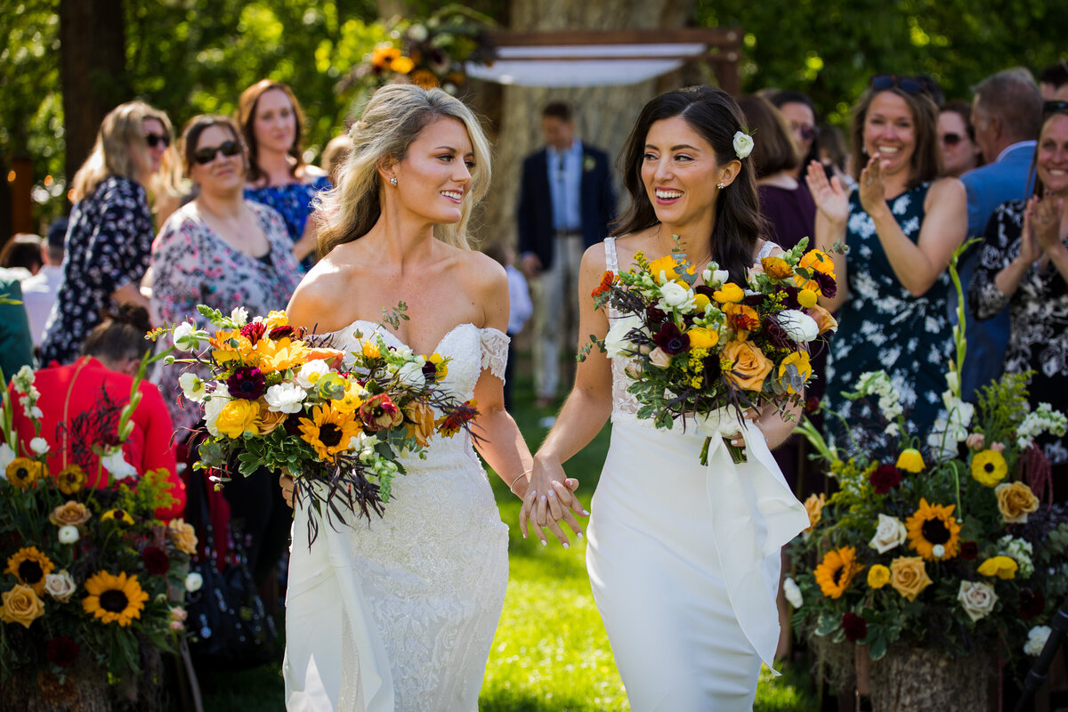 Two brides smile and look at each other as they walk back up the aisle at the end of their ceremony.