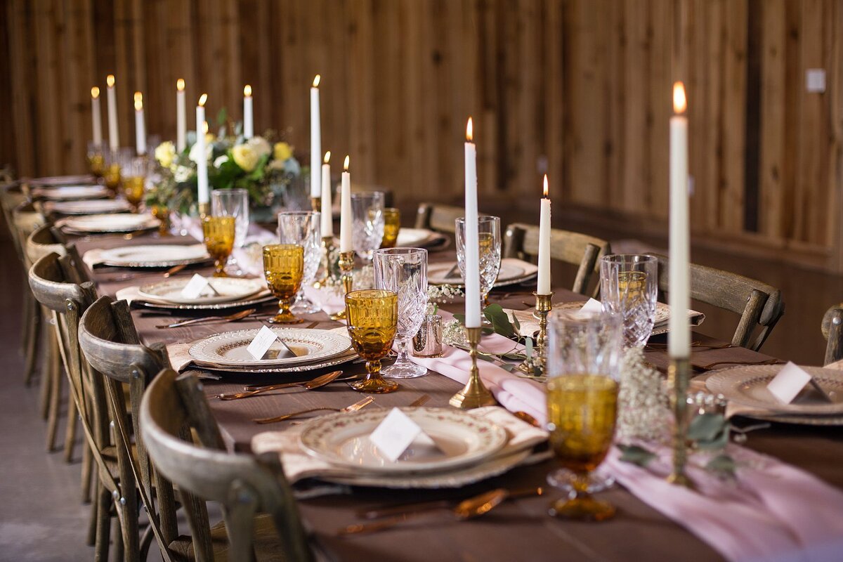 Long dark brown farm table set with ivory chargers and vintage china. Long blush table runners with green garlands and a full centerpiece are in the middle of the table accented with amber crystal goblets, cold candlesticks and white taper candles. Cross-back farm chairs  line the rustic table.