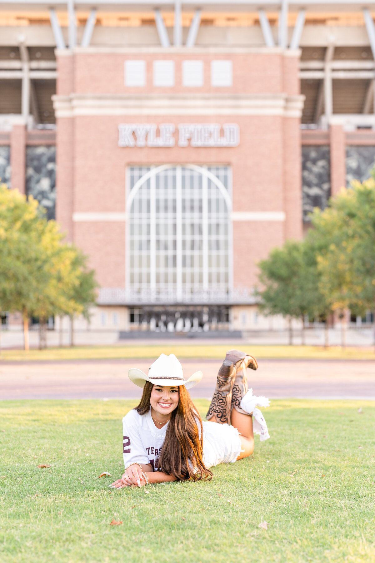 Texas A&M senior girl laying in grass and smiling with feet up while wearing white jersey and cowboy hat in Aggie Park while in front of Kyle Field