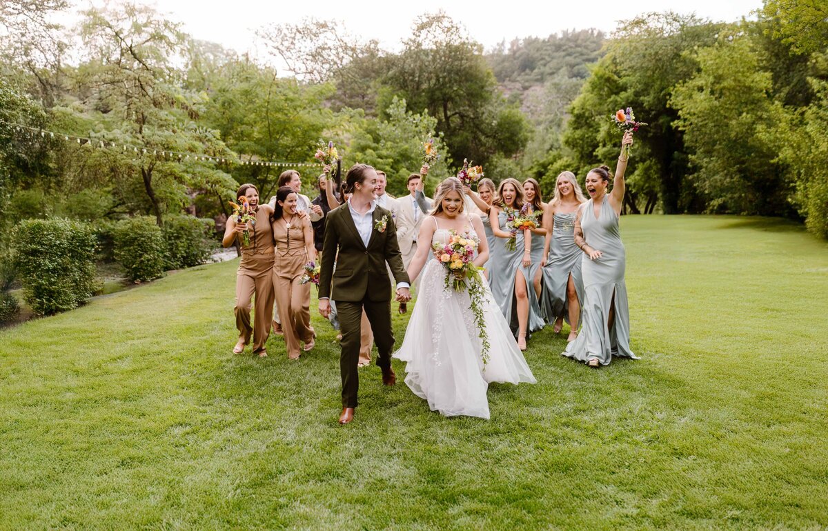 A joyful wedding party celebrates outdoors as the newlyweds walk hand in hand across the lawn, captured by a Colorado Wedding and Portrait Photographer.