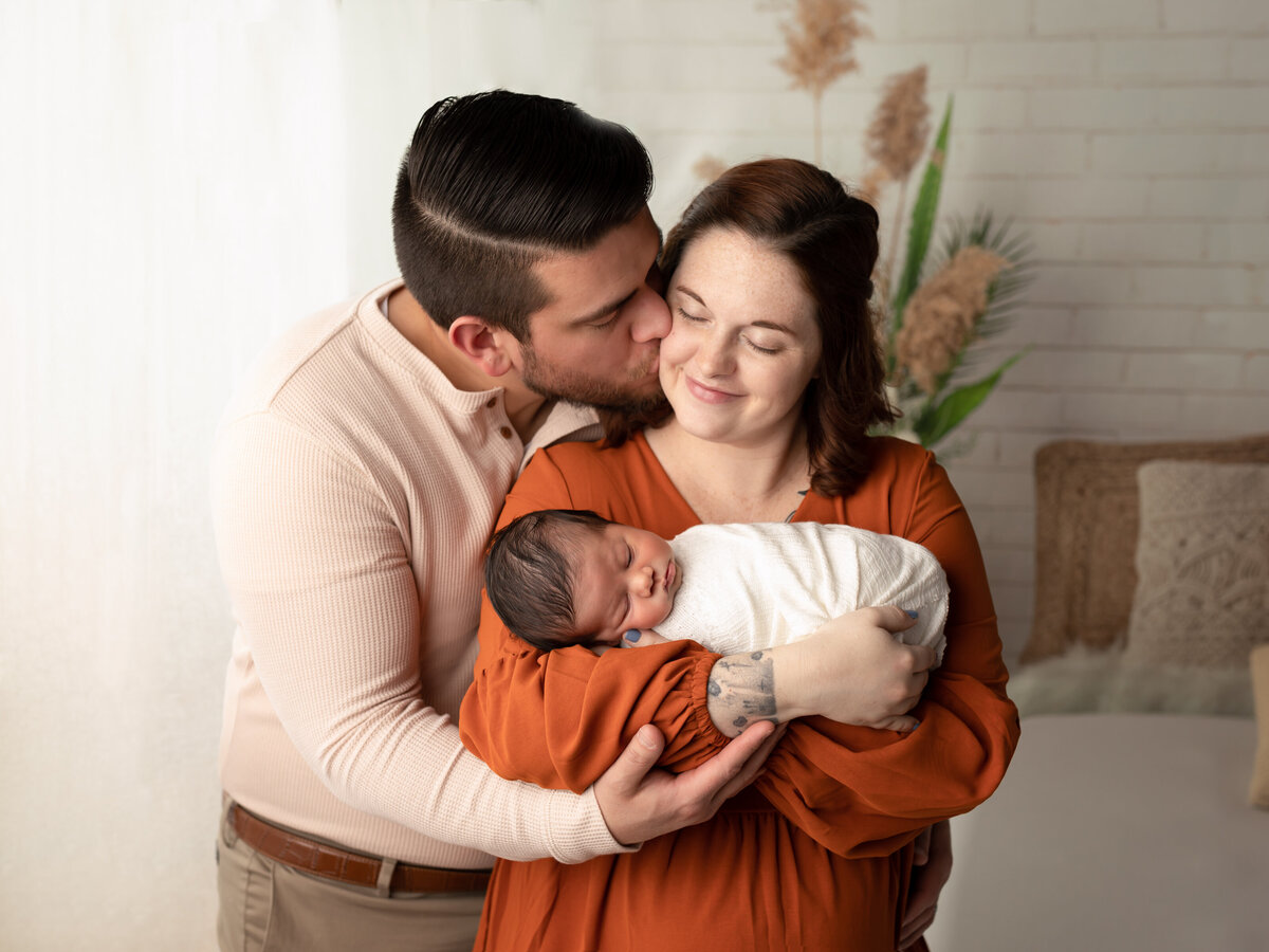 husband kissing wife on the cheek holding newborn baby boy