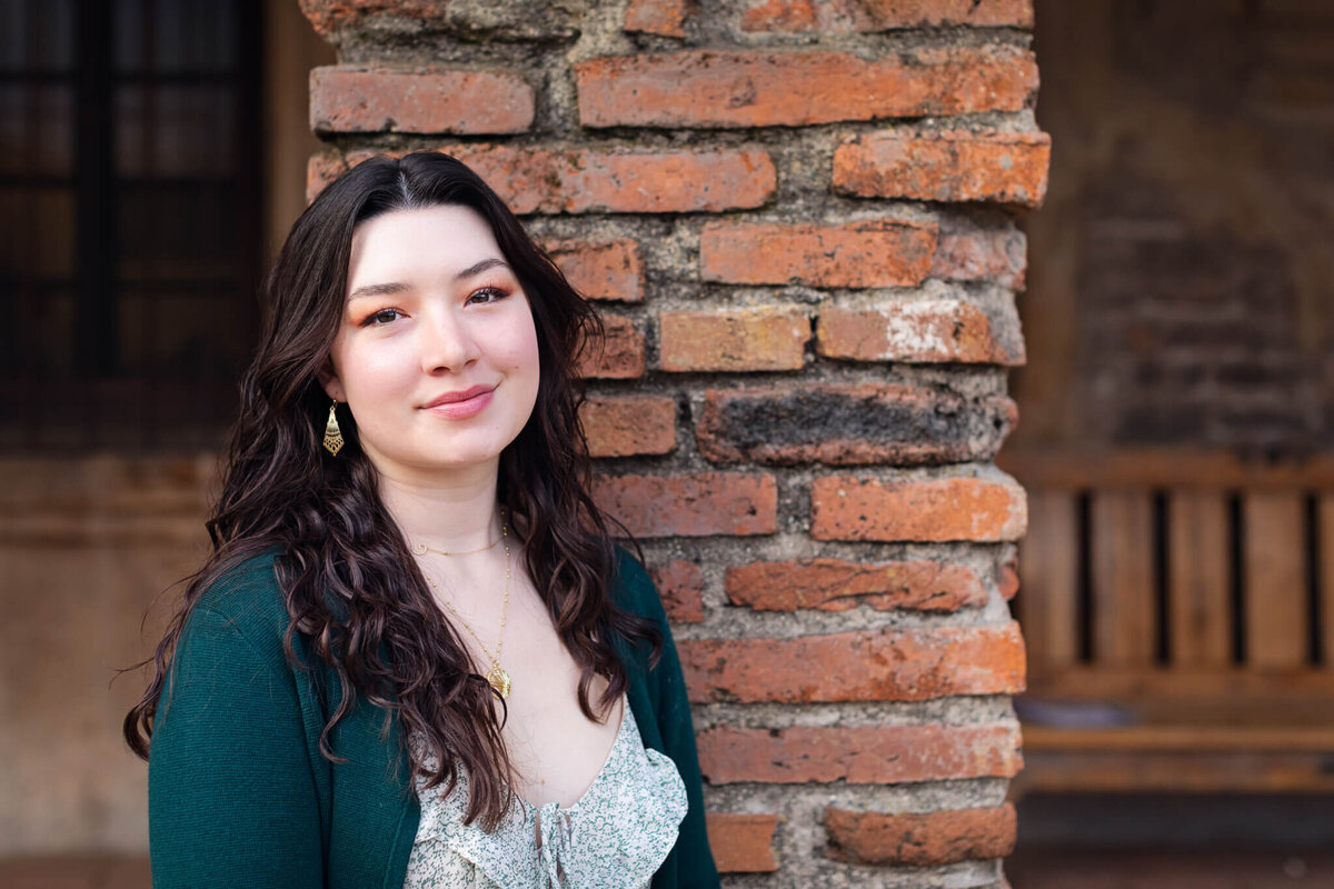 A young woman with dark, wavy hair and a green cardigan smiling softly at the camera in front of a brick column at Mission San Juan Capistrano.