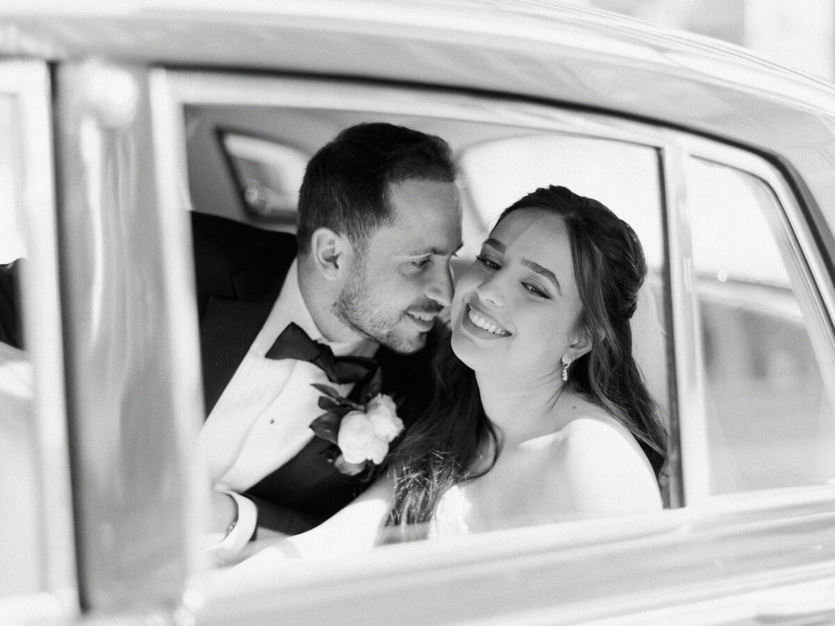 Bride and groom smiling at each other inside a car, captured in a black and white photograph. Their joy perfectly encapsulates the elegance of a classic Calgary wedding.