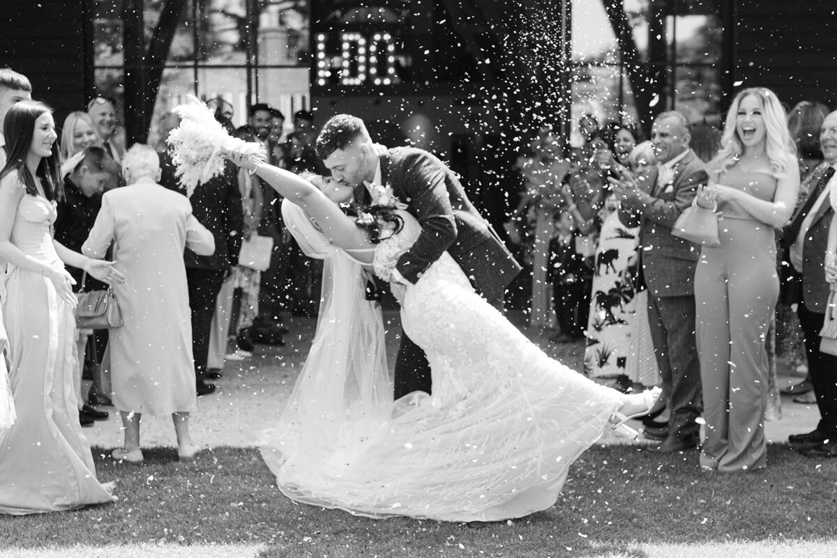 black and white image of a bride and groom kissing during their confetti tunnel. The groom is dipping the bride back for the kiss and they are surrounded by their guests