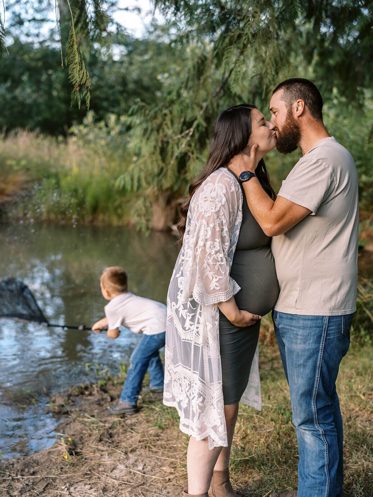 A pregnant woman kisses a man with a boy fishing in the background.