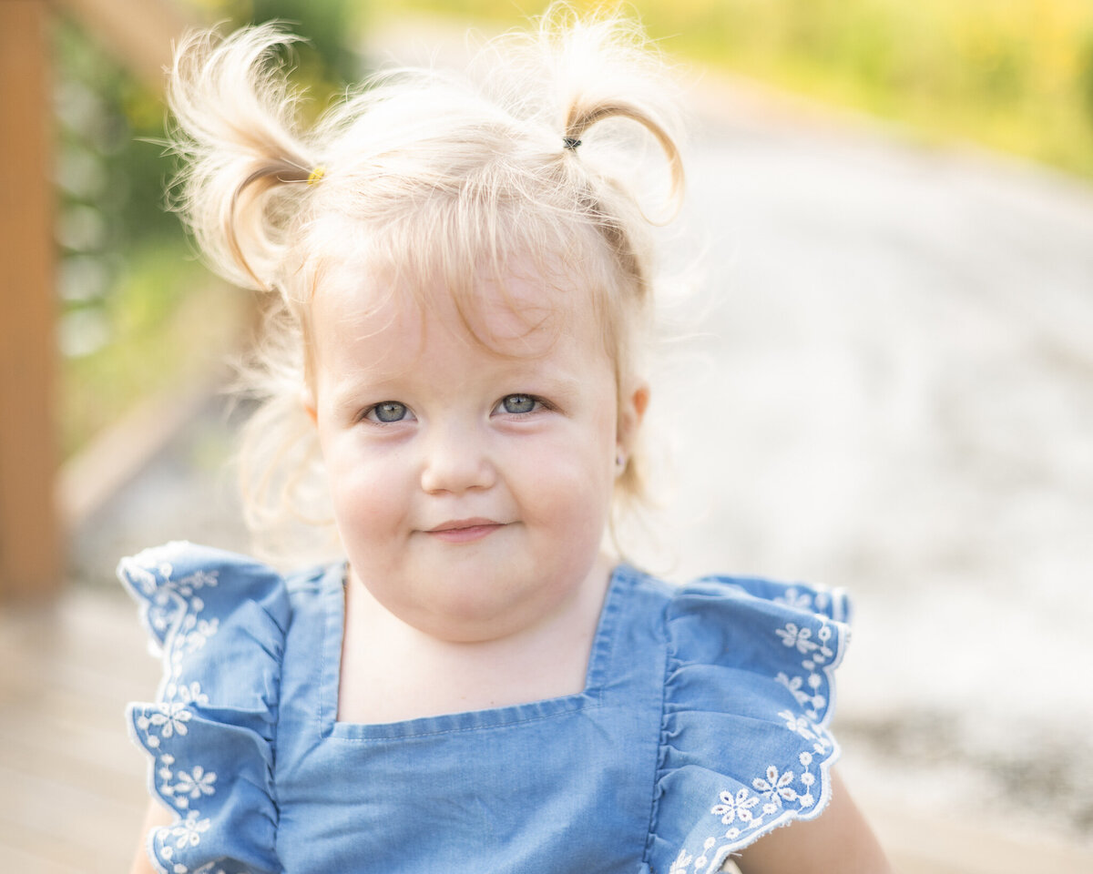 Toddler with light blonde hair in pig tails, standing in a rocked path, wearing a light blue and white dress, smiling at the camera