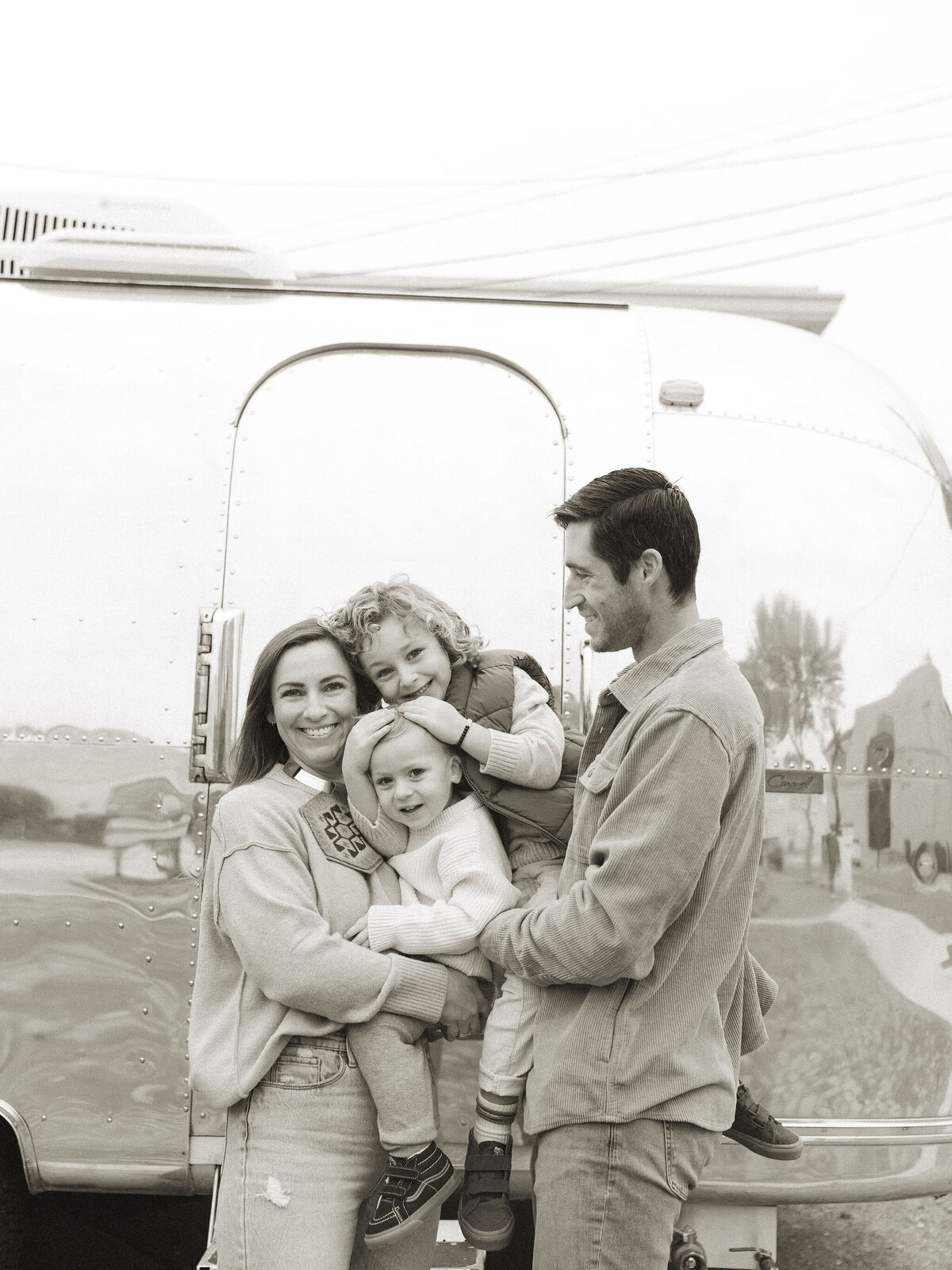 A mother and father hold their two cute boys in front of an airstream trailer. 