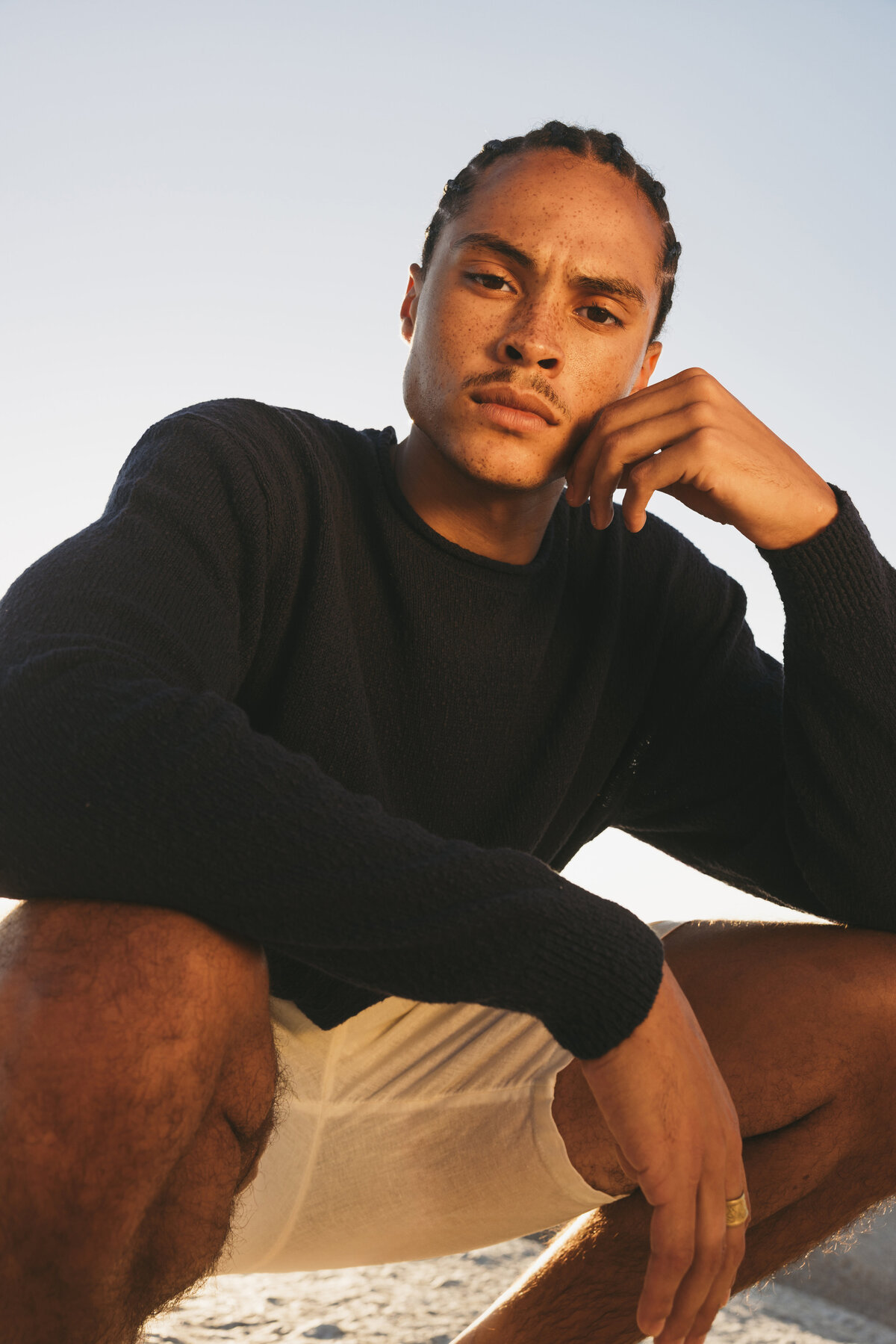 Low-angle close-up of Timothe posing in a dark sweater and white linen shorts, with a serious expression during golden hour in Cape Town.