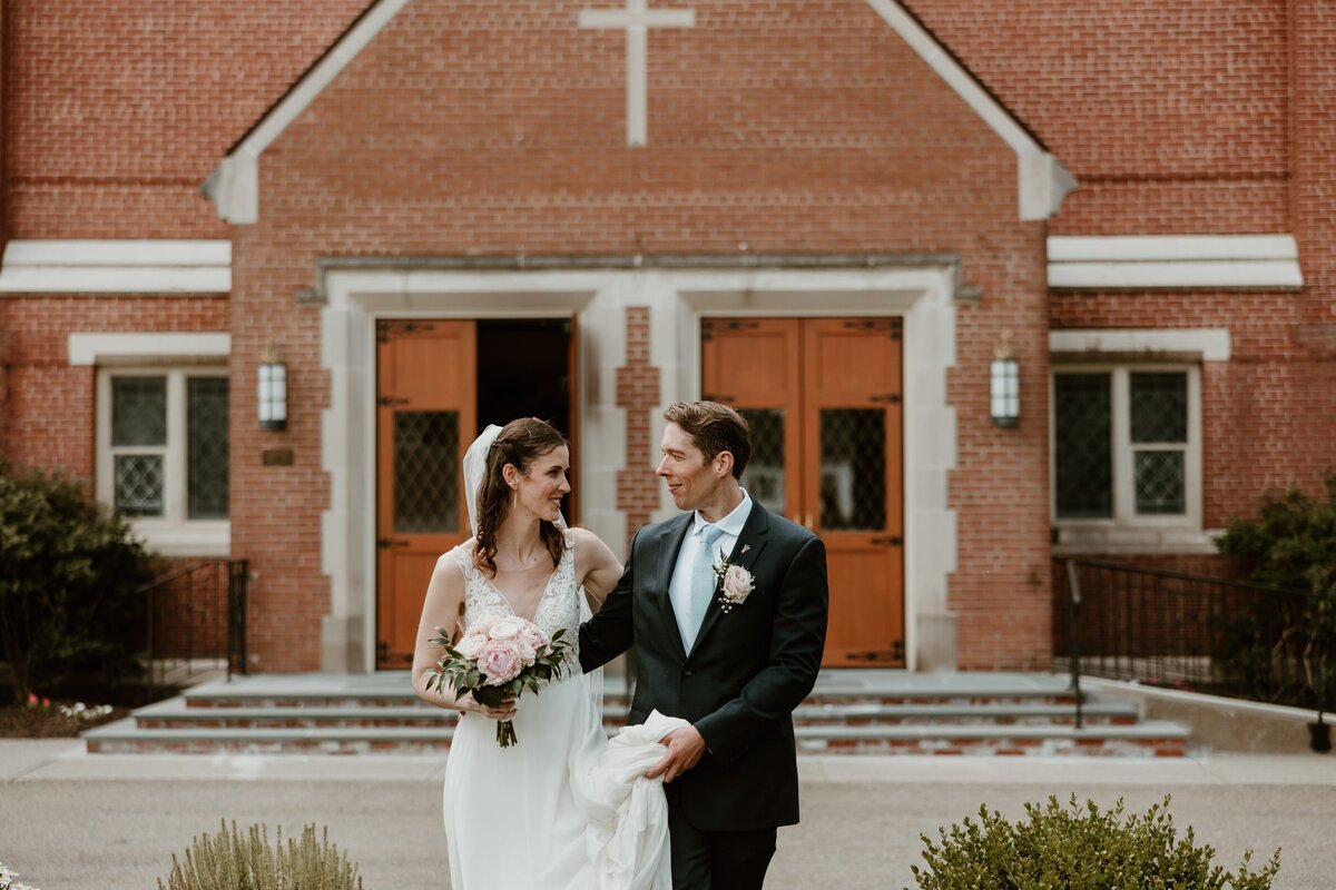 A bride and groom walk arm in arm outside St. Mary's Church in Wappingers Falls. The bride holds a bouquet of pink roses and greenery, smiling lovingly at the groom, who is dressed in a dark suit with a light blue tie and boutonniere. The brick facade of the church and its wooden doors provide a classic and charming backdrop.