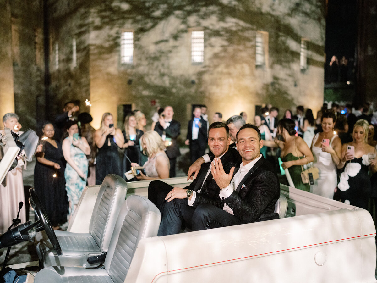 A couple waves joyfully from a classic vehicle celebrating their special occasion, surrounded by friends and family at wedding..