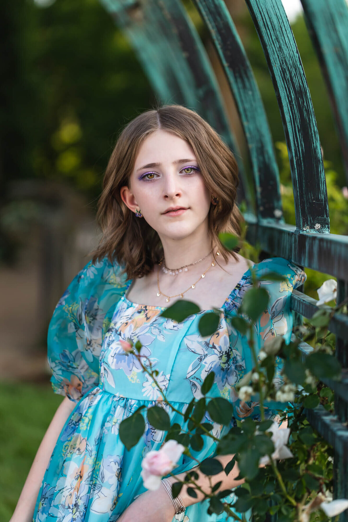 Beautiful green eyed teenage girl in a blue floral dress posing under a rose trellis. Captured by Springfield, MO teen photographer Dynae Levingston.