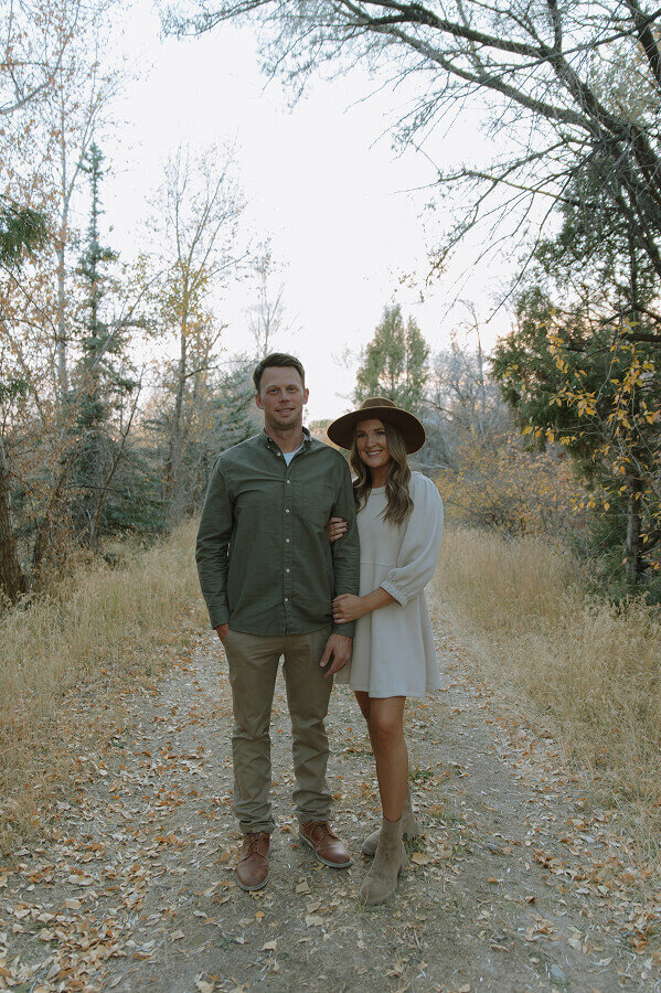 A wife holds her husband's arm as they stand and smile for the camera. They're standing on a dirt road with trees all around them.