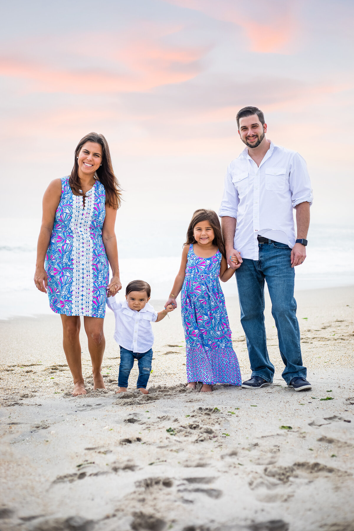 family of four standing in front of ocean in blue and white with pink clouds behind them