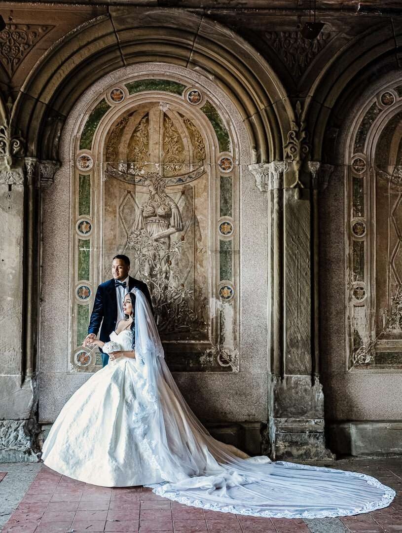 Bride and groom pose in elegant wedding attire under the historic arches at Bethesda Terrace in Central Park, New York City.
