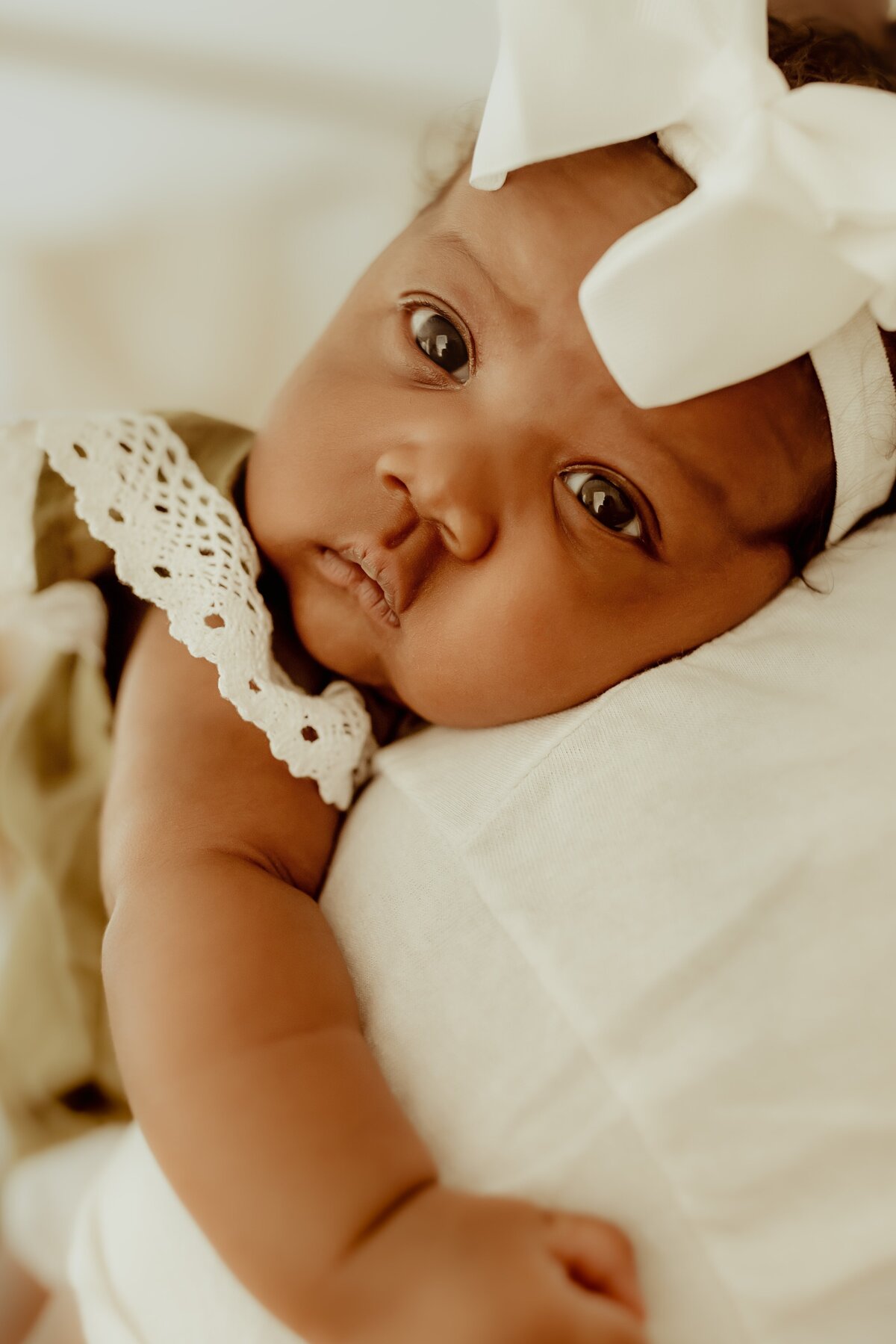 A baby with a white bow headband and wearing a lace-trimmed outfit lies on a soft surface, looking up with wide eyes.