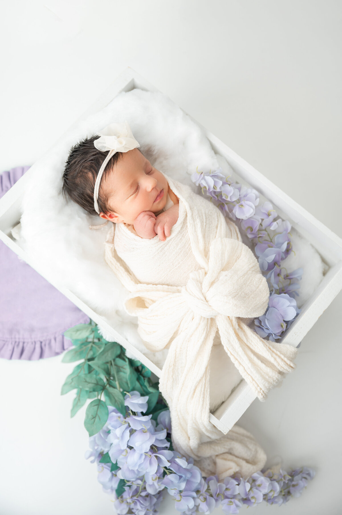 Newborn wrapped with a bow, resting on a white crate framed by lilac details.