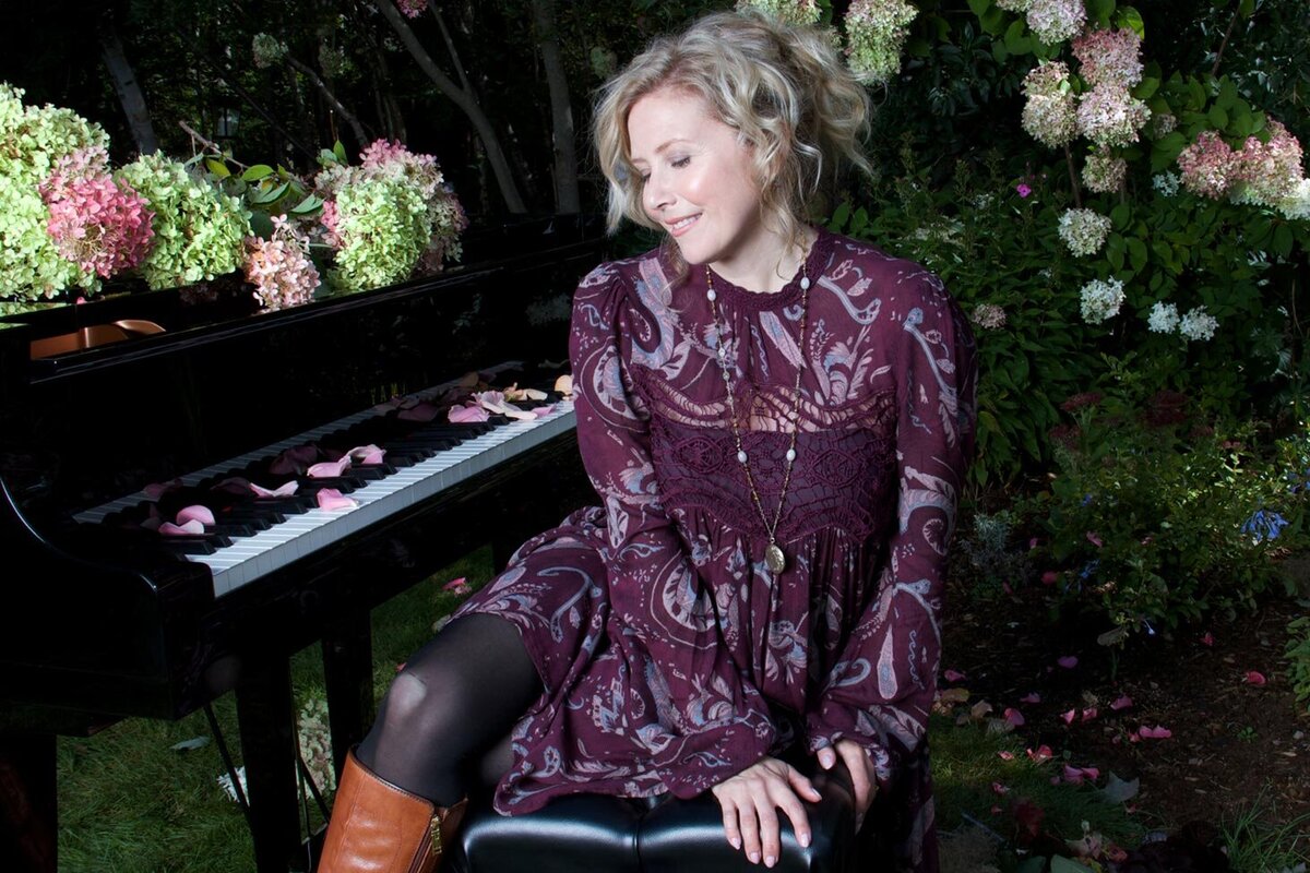 Musician portrait Linda Brooks sitting on piano bench in front of grand piano in garden wearing purple flowered dress