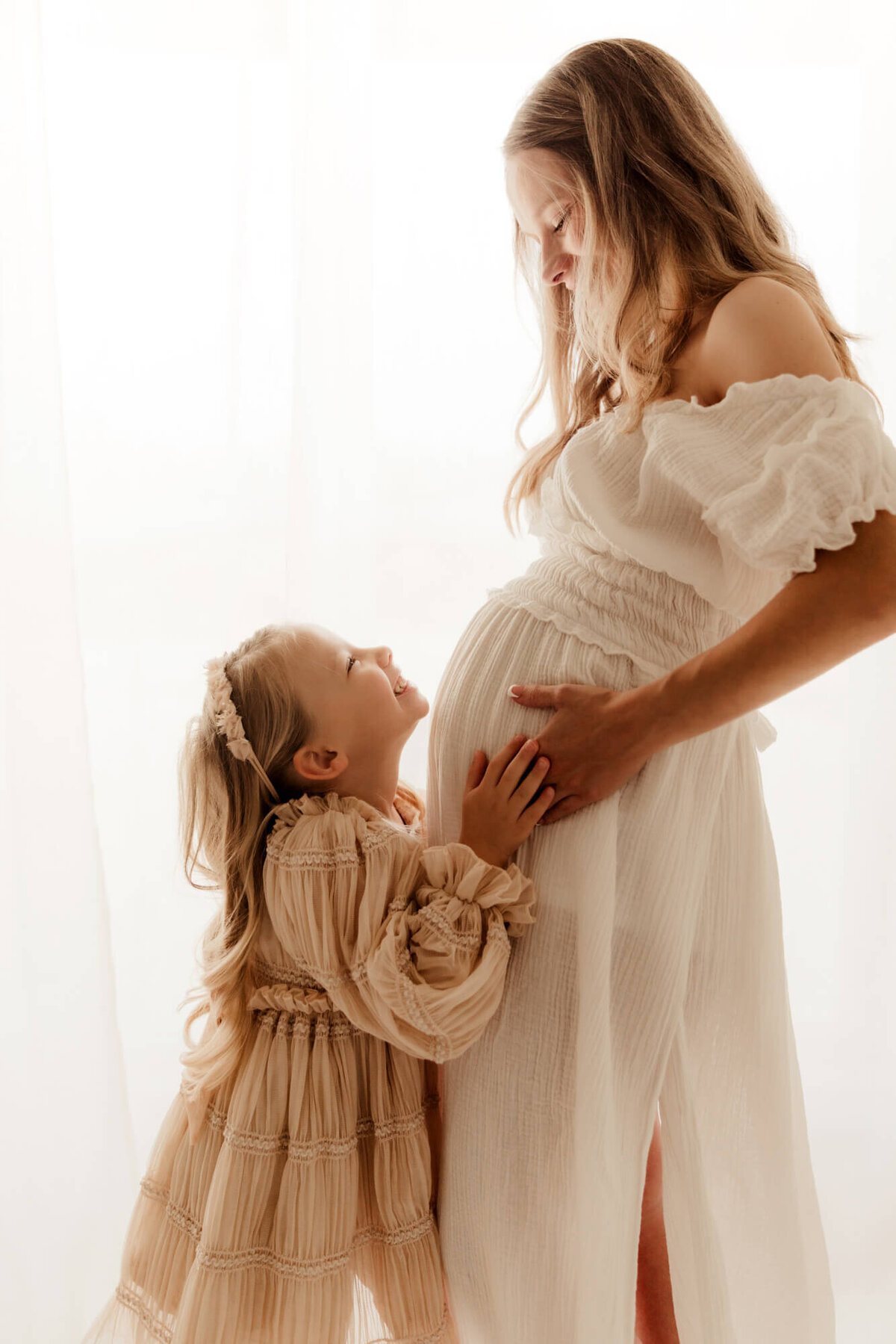Girl wearing a pink dress smiles up at her momma while placing her hands on her mother's pregnant belly in Oklahoma City.