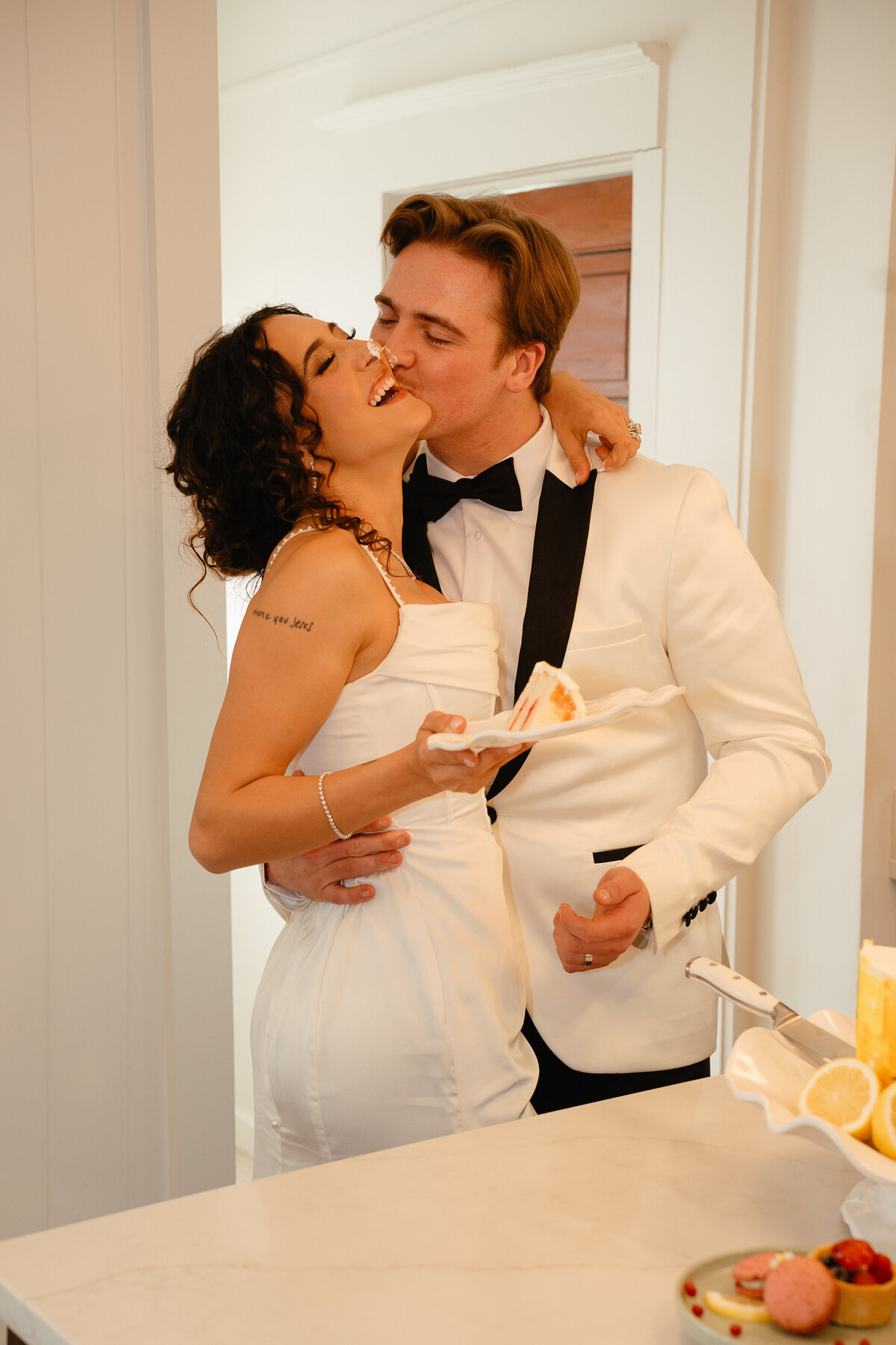 Groom kisses bride on the cheek while she laughs and holds a plate with a piece of cake
