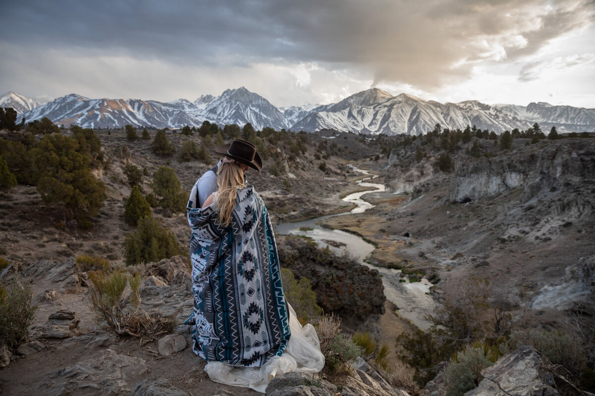 A bride and groom cuddle together under a blanket while they watch the sunset in the snowcapped mountains in Mammoth, California.