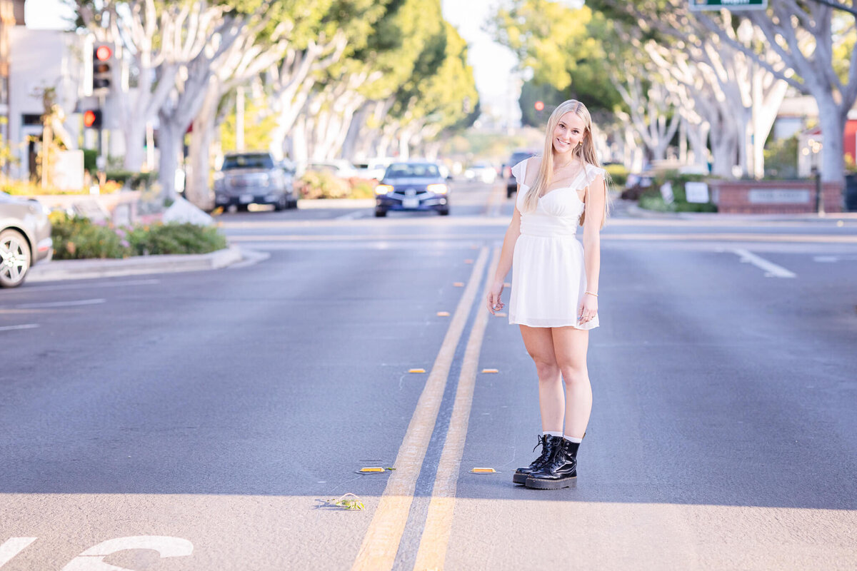 A young woman in a white dress and black boots stands on a sunny street, with cars and trees lining the background.