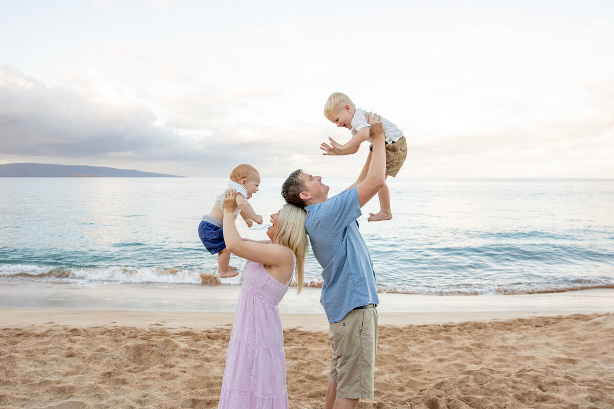 Maui Family Photographers capture woman wearing pink dress lifting child into air and man wearing blue shirt lifting child in air during maui beach family photos