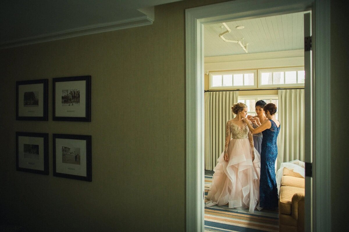 A candid moment of a bride getting ready, assisted by a bridesmaid in a blue dress, viewed from the doorway of a warmly lit room.
