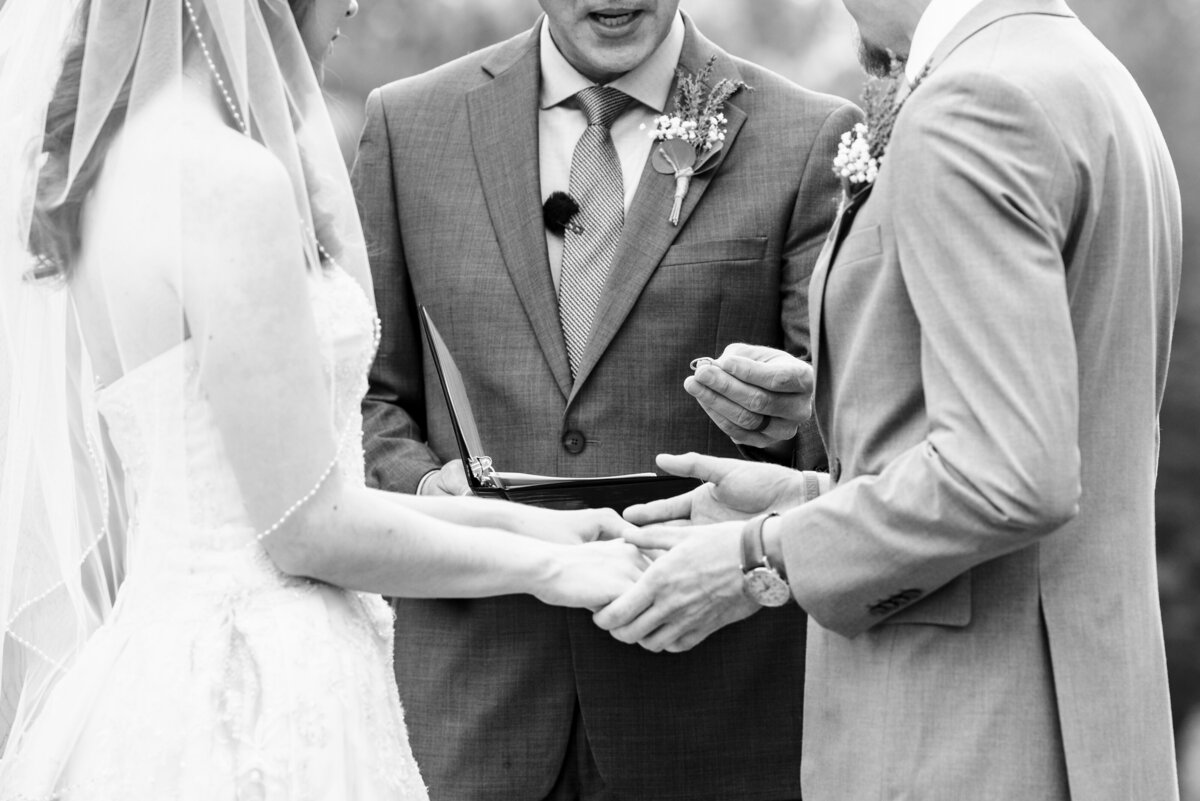 Tight framed black and white photograph of officiant holding out a wedding band to a groom by Charlotte wedding photographers DeLong Photography