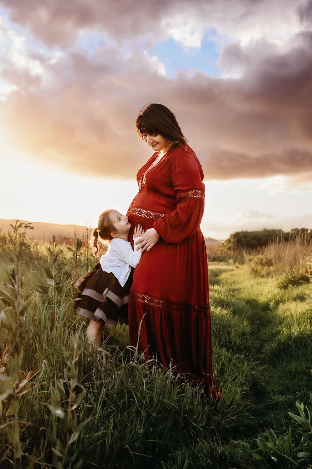 girl looks up at pregnant mama. mom wears a red maternity dress