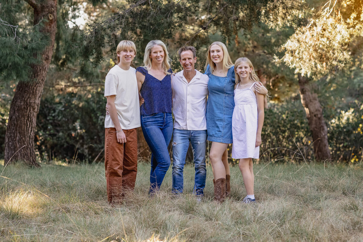 A family of five with blonde hair wearing shades of blue, white, and brown, standing in the grass in front of many trees, smiling at the camera.