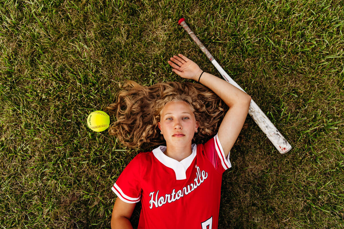 Gorgeous high school senior laying in the grass with her softball and bat during near Green Bay
