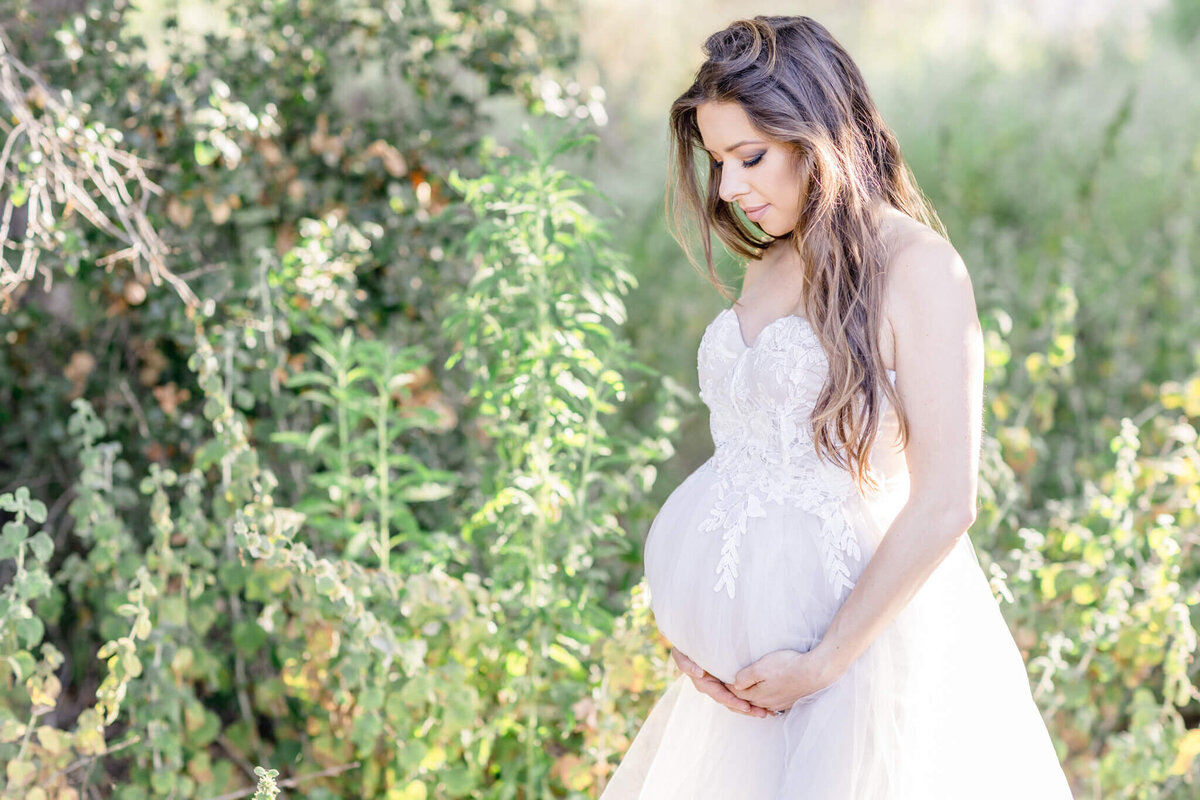 A pregnant woman in a white lace dress stands in a sunlit garden, gently holding her belly. Her long hair cascades over her shoulders as she looks down with a serene expression. Lush green foliage surrounds her.